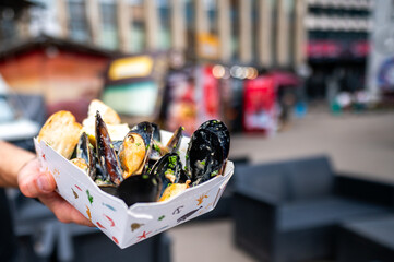 A hand holding a white paper container filled with freshly cooked mussels, garnished with herbs,...