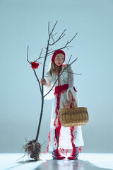 Young woman in white lace dress, red accents, and knitted hat holding basket and standing beside...