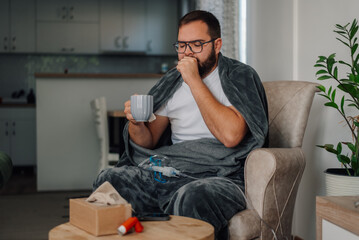 Fototapeta premium Sick man coughing at home, drinking tea and using inhaler