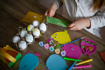A child is making crafts from felt and paper on a wooden table. Nearby are decorative eggs, googly...