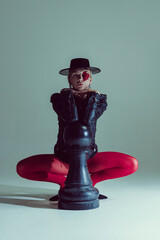 Seated woman with red eye patch, hat, and structured black jacket resting on oversized chess piece,...