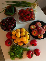 Fresh fruits and vegetables lying on a white table in the sunset light from the window. Apricots, tomatoes, parsley, green onions, strawberries, cherries, cucumbers and peaches
