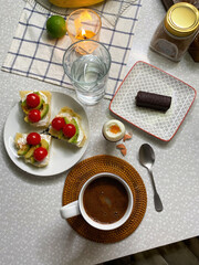 Flat lay breakfast with a large mug of coffee, avocado and cherry tomato sandwiches. Chocolate cheese on a plate and a scrambled egg, candle burning for atmosphere. Cozy serving homemade breakfast