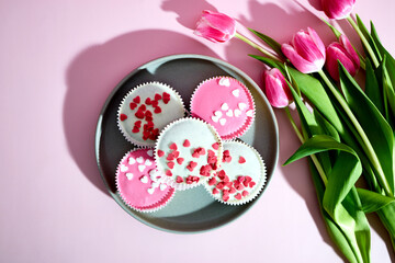 Sponge cupcakes with pink and white icing and heart-shaped sugar sprinkles on a pink background next to tulip flowers