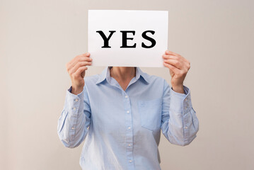 Woman holding a white card with the word yes written on it, covering her face on beige background