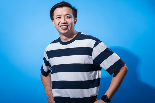 Confident Asian man smiles while posing with hands on hips, wearing a black and white striped shirt against a clean blue background in a brightly lit studio setup for portraits