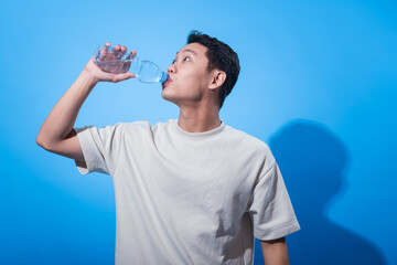 Young Asian man in a plain cream t-shirt drinks from a clear plastic water bottle while looking up, standing against a solid blue background, promoting hydration and a healthy lifestyle