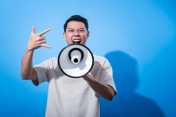 Asian man in a cream t-shirt shouting through a megaphone while showing a thumbs-up gesture, expressing approval and excitement against a bright blue studio background