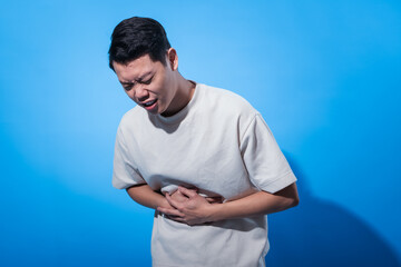Asian man in a plain white t-shirt clutches his stomach with both hands and screams in pain, eyes...