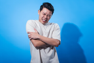 Asian man in a white t-shirt holding his arm tightly with a painful facial expression and eyes...