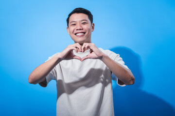 Smiling young Asian man in beige t-shirt making a heart shape with his hands in front of chest against a blue background, expressing love, kindness, and positivity in a cheerful studio portrait