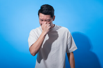 Asian man in a white t-shirt holding his nose and squinting his eyes in discomfort against a bright...