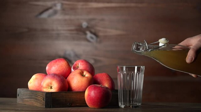 Man pouring apple cider from a glass bottle into a glass next to fresh red apples in wooden tray on kitchen table, close up