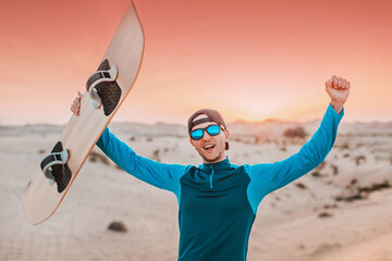 Young male athlete is raising arms and holding sandboard after sandboarding training in the desert at sunset