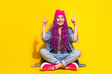 Trendy young woman with vibrant hairstyle in denim attire pointing upwards, sitting cross-legged on a yellow background.
