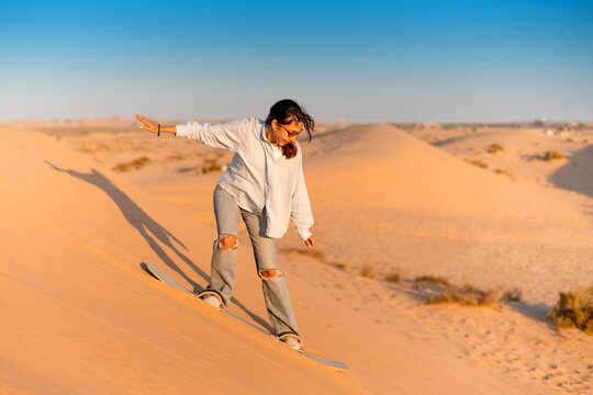 Young woman sandboarding down a sandy dune in the desert, enjoying the thrill of the sport during a stunning sunset