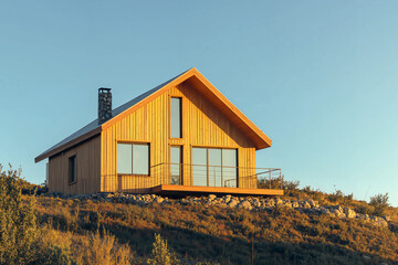 Obraz premium A modern wooden house with large windows, a balcony, and a scenic mountain background during golden hour.