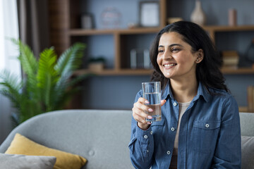 Photo of an Indian young woman in casual clothes at home, sitting on the couch, holding a glass of...
