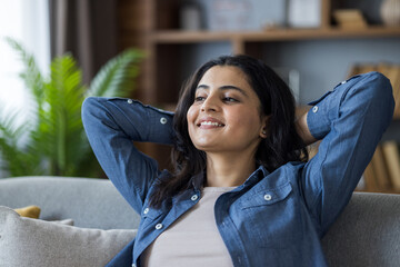 A close-up photo of a young Indian woman sitting on a couch, arms behind her head, leaning to the...