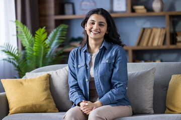 Portrait of a smiling young Indian woman in casual clothes and denim shirt sitting on the couch at...