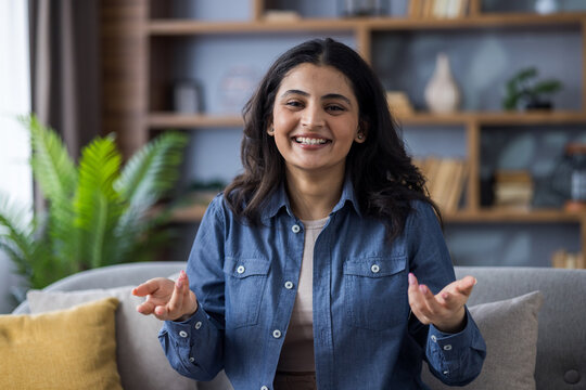 Close-up portrait of a young Indian woman in a denim shirt sitting on a sofa at home in front of the camera and talking while gesturing with her hands