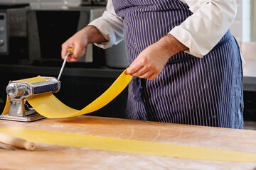 Italian traditional restaurant chef rolling out dough on a metal dough sheeter machine for homemade pasta on wooden table 
