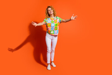 Happy young woman with red hair in casual floral outfit posing against vibrant orange background expressing joy through open arms