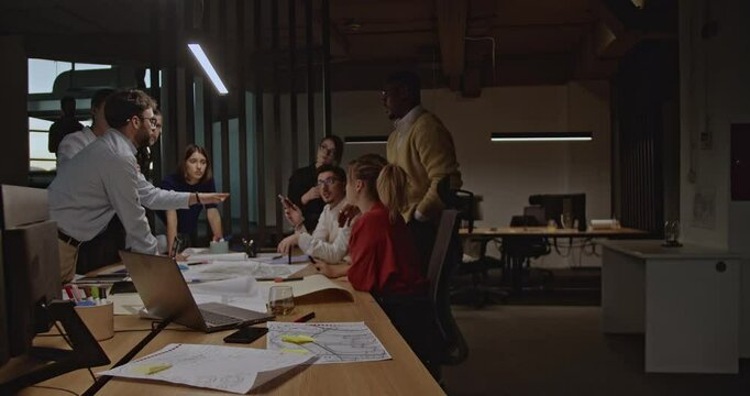 A diverse team of business professionals collaborates on a project in a modern office, reviewing blueprints and engaging in discussions.