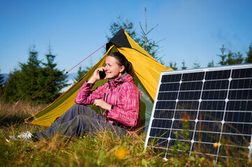 Happy woman sits in front of yellow tent, talking on her phone. Solar panel, highlighting use of...