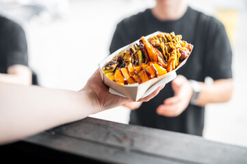 A hand passes a tray with grilled and fried food, drizzled in sauces, showcasing a street food...