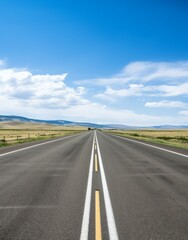 Fototapeta premium Endless two-lane highway stretching into the horizon through a vast rural landscape under a bright blue sky with scattered clouds.