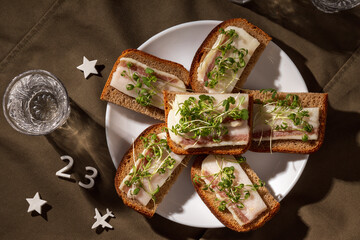 A beautifully styled food scene featuring rye bread with salo and microgreens, set on a textured cloth with a vodka glass and decorative number '23'. 