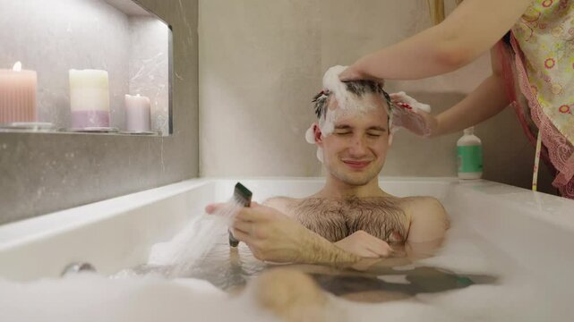 A young man relaxes in a bubble bath while his girlfriend massages shampoo into his hair, creating a calming and intimate moment.