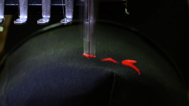 Machine embroidery of a black hat. Embroidering a design with red thread shot close-up. Concept of: Automated work, Embroidery machine, Thread, Fabric, Needle, Hat. 4k video 50fps.