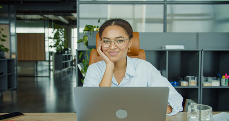 A smiling young woman with glasses sitting at a desk and working on her laptop in a modern, bright...