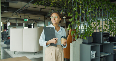 Confident young woman in a stylish office holding a clipboard and coffee cup, walking with a smile in a modern workspace featuring green plants and contemporary furniture