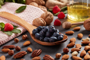Une cuillère en bois pleine de myrtilles repose sur une surface grise parsemée de noix, d'amandes, de noisettes, de framboises et d'un pot de miel.