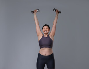 Plus size woman in gym clothes doing exercise with dumbbells on grey background