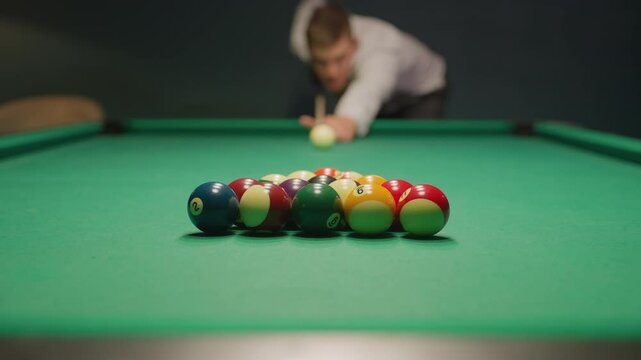 Blurred figure in light shirt and dark trousers leans over green pool table, gripping cue stick with deep concentration. Eyes locked on balls, posture steady, creating tension in dimly lit game room