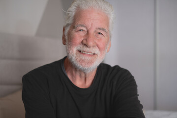 Portrait of an handsome old smiling caucasian senior bearded man looking at camera, sitting inside home