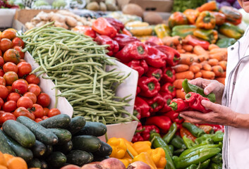 Senior woman's hand holding red and green peppers in hand while buying fresh vegetables at the market choosing from a large selection. Healthy eating, the concept of spending and consumerism