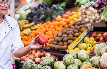 Senior woman holding a red tomato in hand while buying fresh fruit and vegetables at the market choosing from a large selection. Healthy eating, the concept of spending and consumerism
