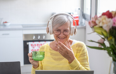 Smiling active modern grey haired senior woman with headphones sitting at home table using laptop...