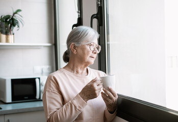 Happy middle aged senior retired woman standing near window, looking in distance, recollecting good...