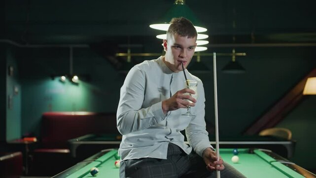 Billiard player in white shirt and grey trousers sits on green pool table holding cue stick and sipping lemon drink. Pool hall setting with stylish overhead lighting and relaxed atmosphere
