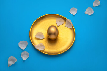 Luxurious golden easter egg rests on a golden plate adorned with delicate white petals. Creating a sophisticated easter scene against a vibrant blue backdrop with water droplets. Viewed from above
