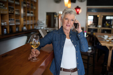 Mature woman enjoying wine while talking on smartphone at cozy bar, copy space