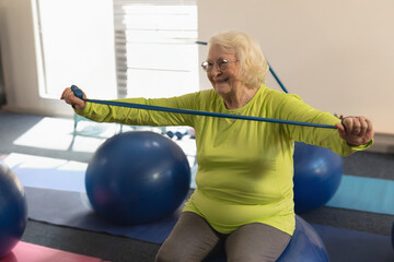 Senior woman exercising with resistance band on yoga ball, smiling in fitness class