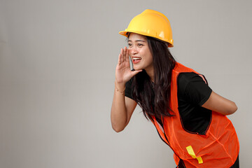 Smiling Asian woman in a yellow hard hat and orange safety vest shouts or calls out with an...