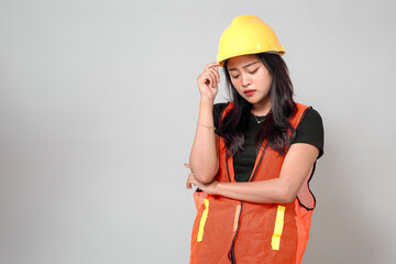 An Asian woman in a yellow hard hat and orange safety vest looks down, appearing stressed and...